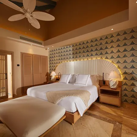 Bahia Principe Luxury Cayo Levantado - All Inclusive Samaná Camas foto The photo shows a luxurious bedroom with a tropical and elegant vibe. The focal point is a large bed with a curved, white, sunburst headboard adorned with vertical slats. The bed is dressed in crisp white linens, accented by a light beige throw blank