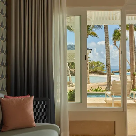 Bahia Principe Luxury Cayo Levantado - All Inclusive Samaná Habitación foto The photo shows a cozy corner of a room with a view of a beautiful outdoor area. 

On the left, a light gray sofa is partially visible. It's adorned with a rectangular coral-pink pillow and a gray patterned wall behind it. A dark, lattice-like screen